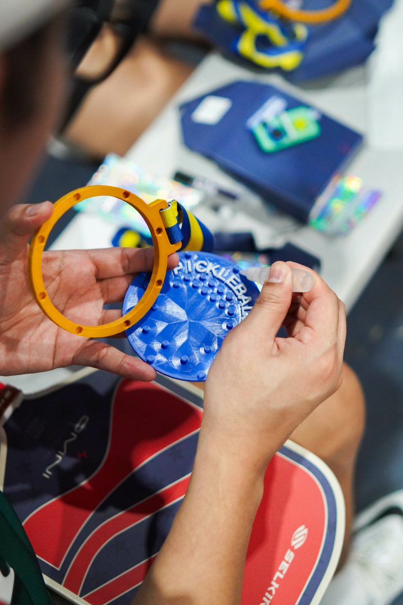 Two hands fitting a yellow printed ring onto a blue medal face, assembling a multi-part 3D printed award by hand.